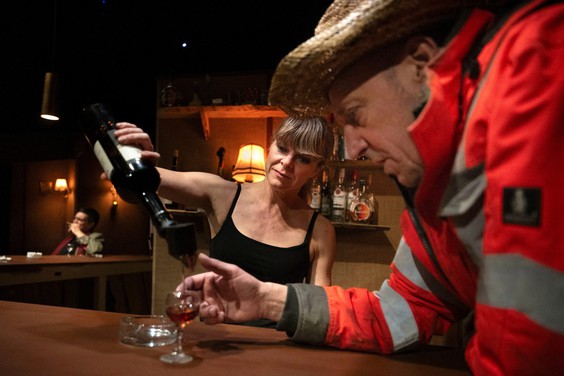 Billede af en julebod. Kvinde med sort jakke, briller og nissehue rækker en juledekoration over bordet til et par.