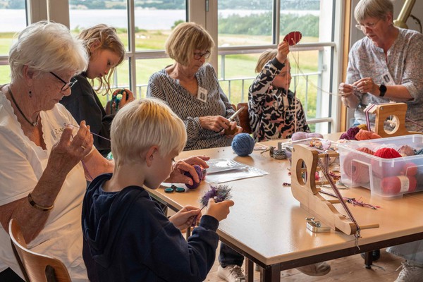 Børn og ældre kvinder sidder om et bord sammen og laver håndarbejde