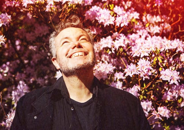 Billede af Rasmus Nøhr. Kigger smilende op i luften og stor foran en lyserød blomsterbusk. Der er et romantisk skær på billedet. 